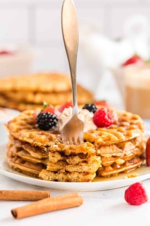 A fork stands upright stuck in pieces of waffle on a plate.