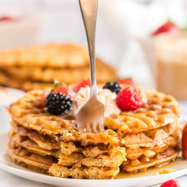 A fork stands upright stuck in pieces of waffle on a plate.