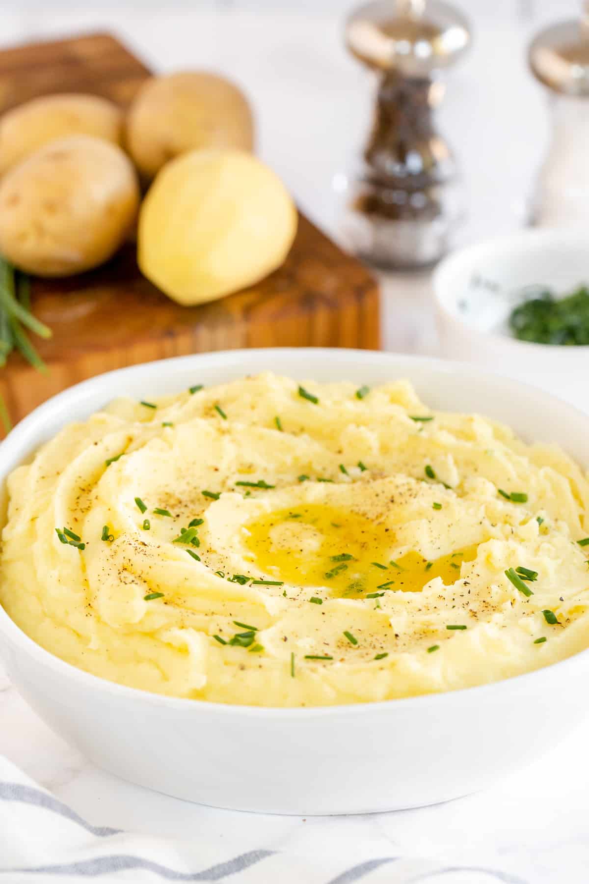 A bowl of mashed potatoes in front of a cutting board with ingredients