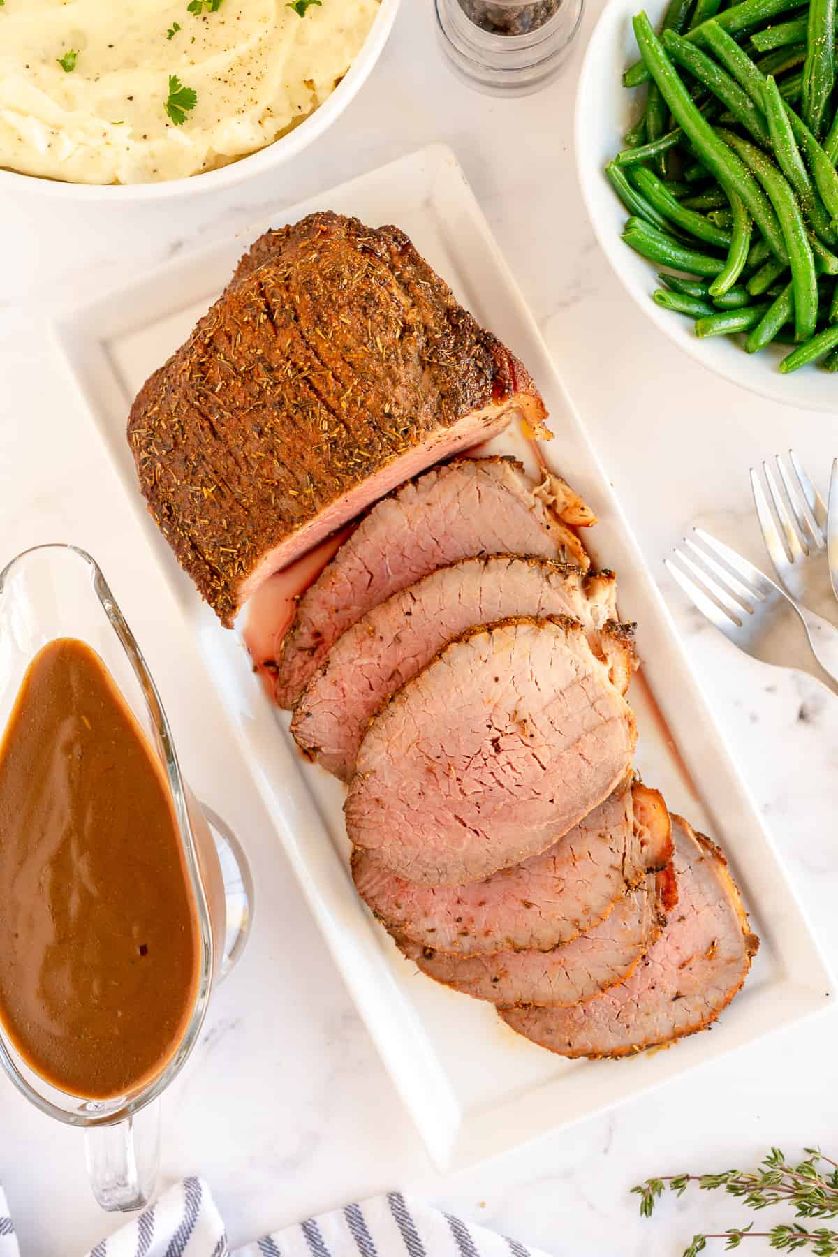 Tender sliced roast beef on a platter next to a gravy boat.