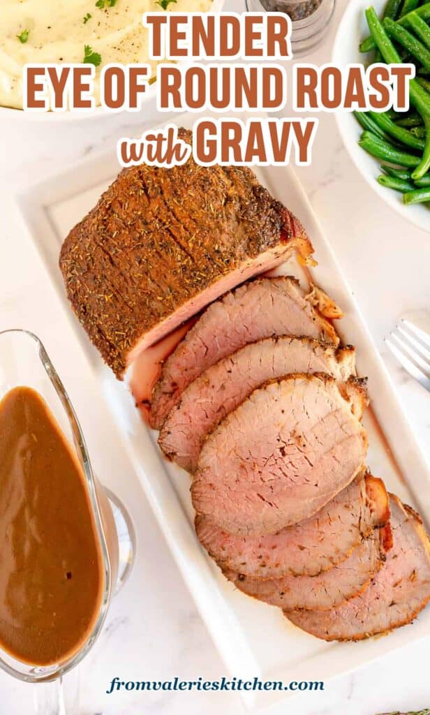 A sliced eye of round roast on a white platter next to a gravy boat with text.