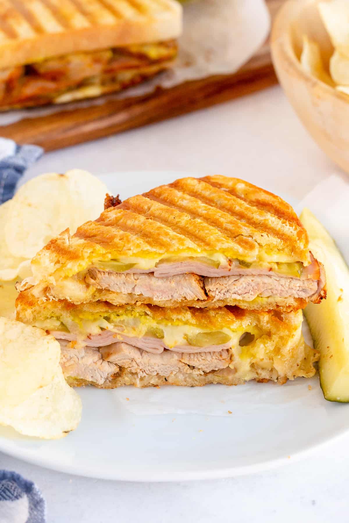 A cuban sandwich on a plate with a pickle and potato chips.