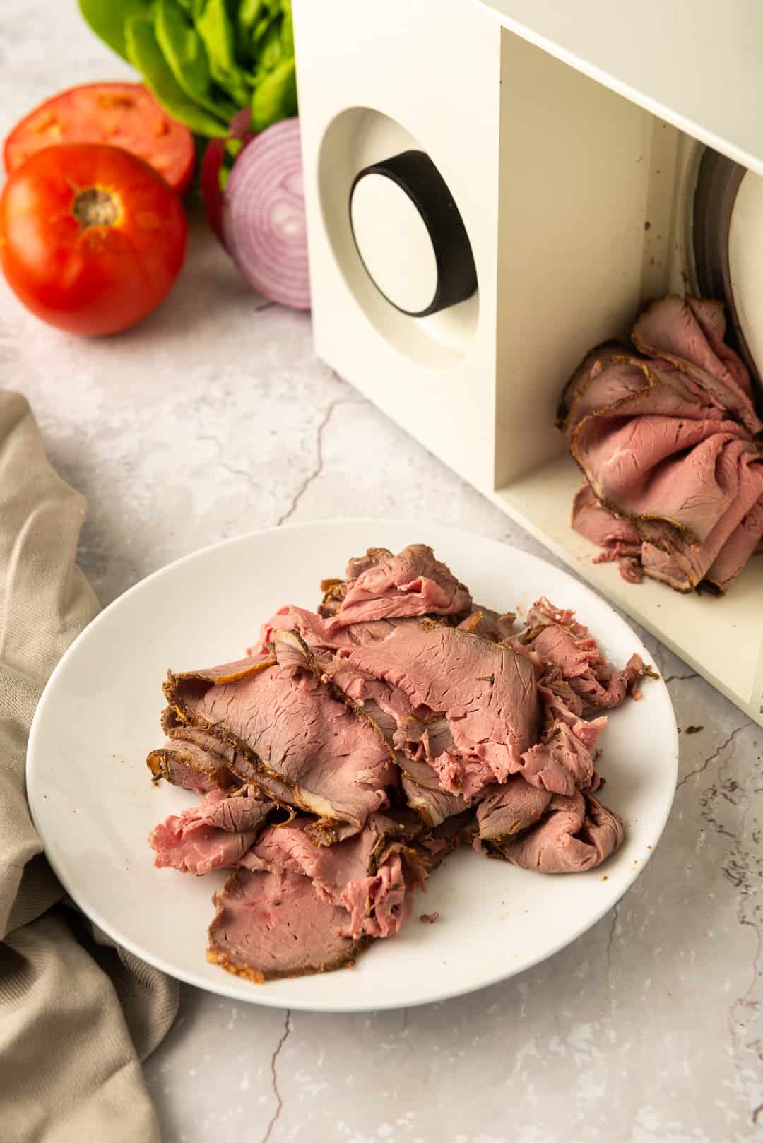 Thinly sliced roast beef on a plate in front of a meat slicer. 