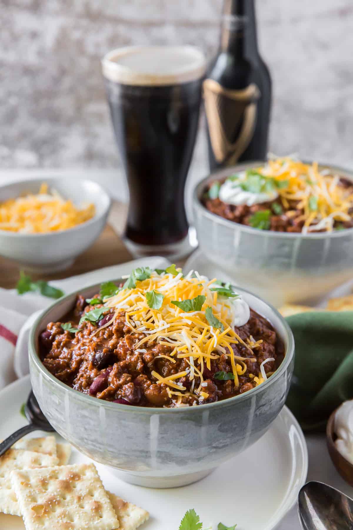 A bowl of chili with a glass of beer in the background.