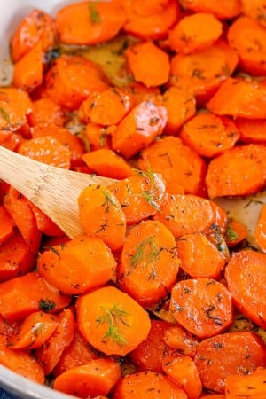 A close up of carrots with dill in a skillet with a wooden spoon.