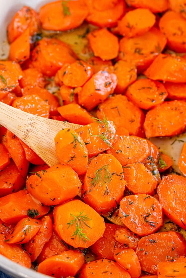 A close up of carrots with dill in a skillet with a wooden spoon.