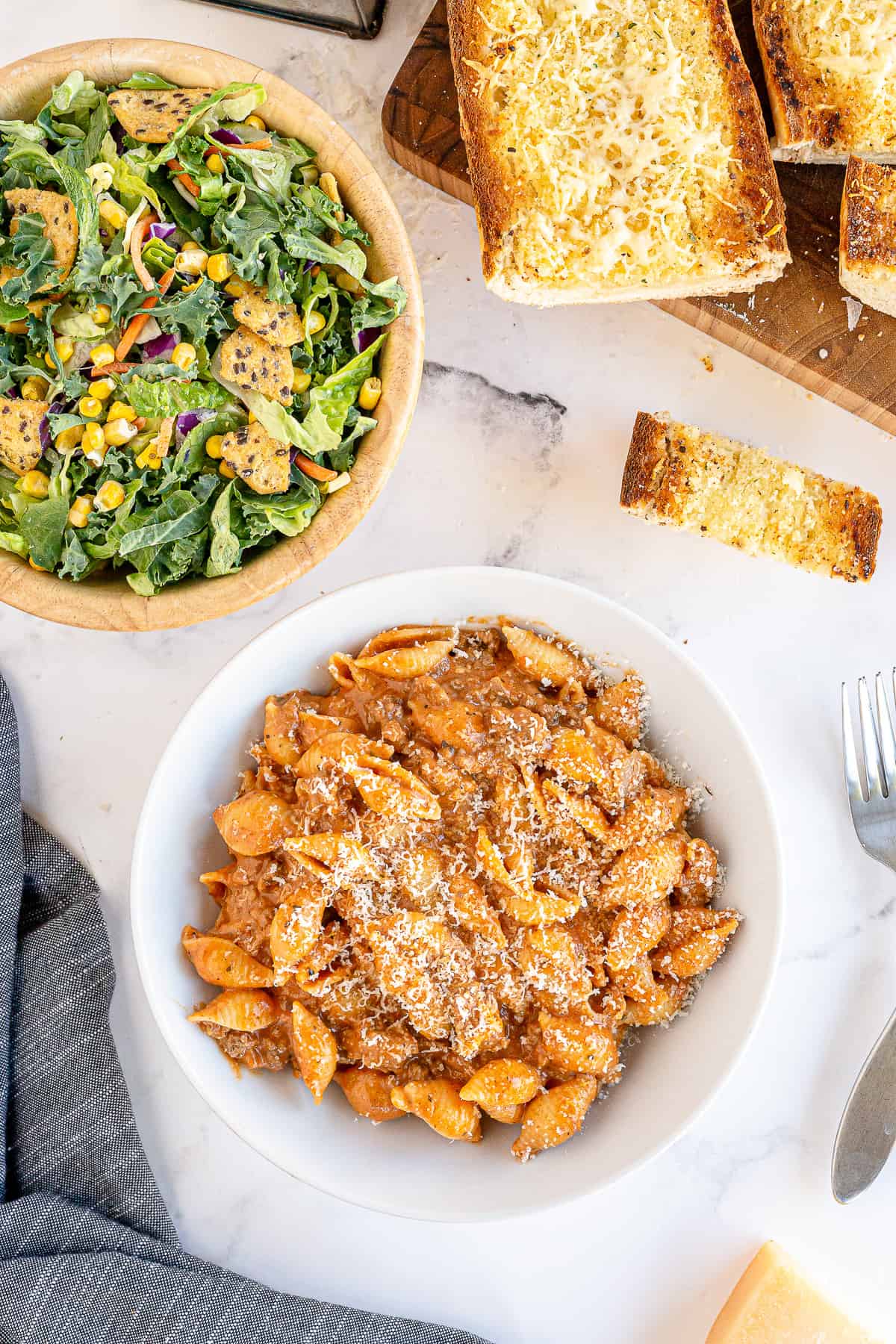 A bowl of pasta next to a big salad and garlic bread.