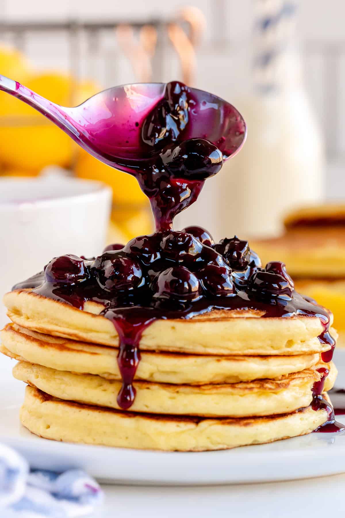 A spoon pours Blueberry Sauce over a stack of pancakes.