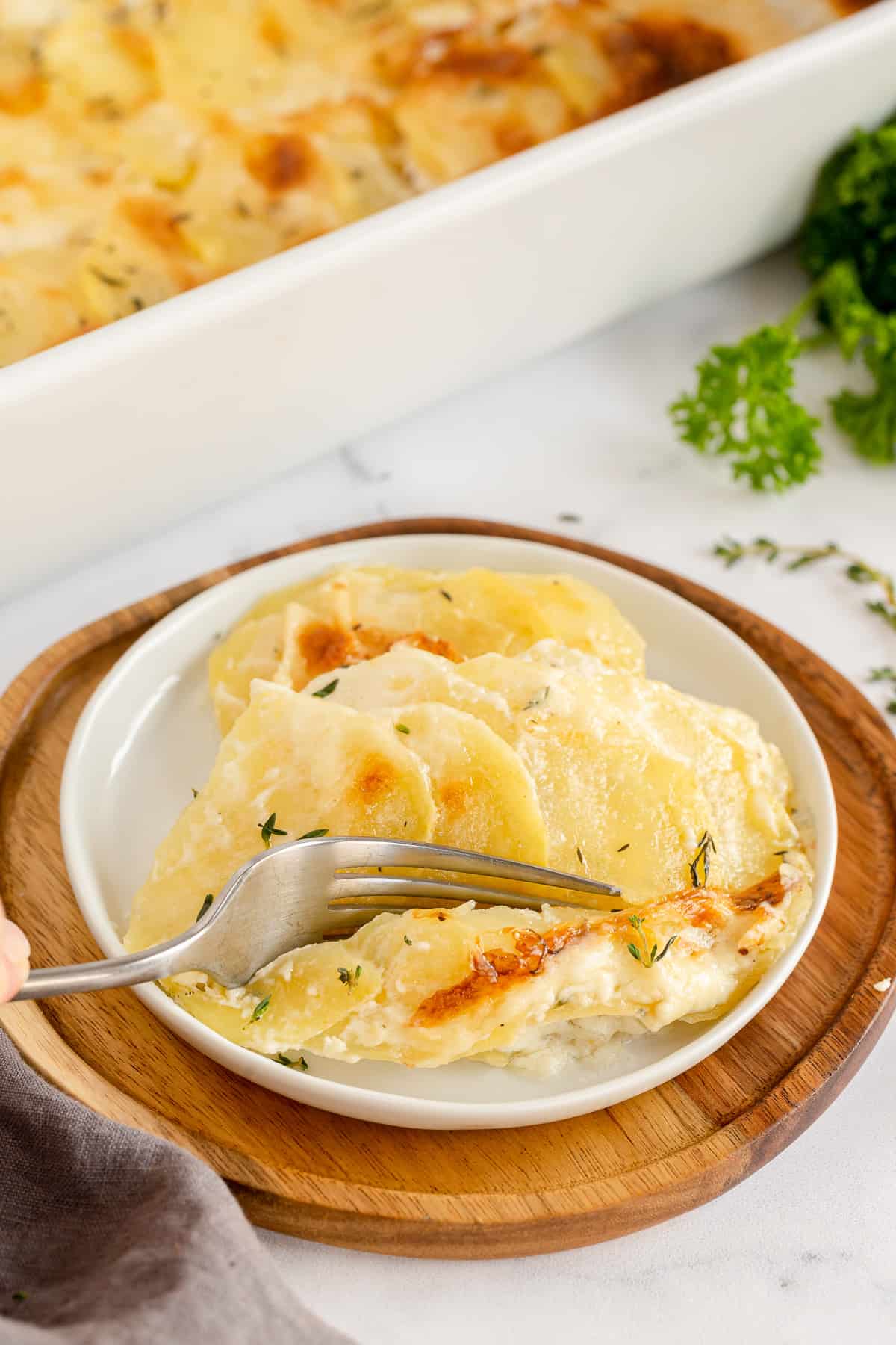 A fork breaks into a serving of scalloped potatoes on a small white plate.