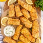 Fish sticks on a cutting board with tartar sauce and lemon wedges.