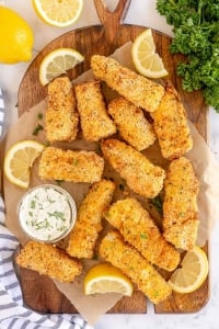 Fish sticks on a cutting board with tartar sauce and lemon wedges.