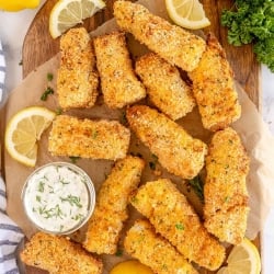 Fish sticks on a cutting board with tartar sauce and lemon wedges.