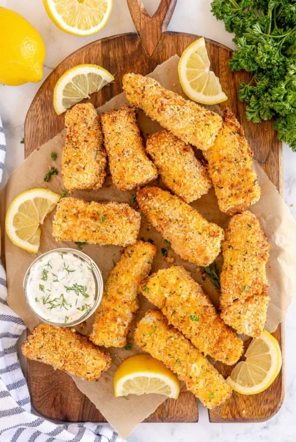 Fish sticks on a cutting board with tartar sauce and lemon wedges.