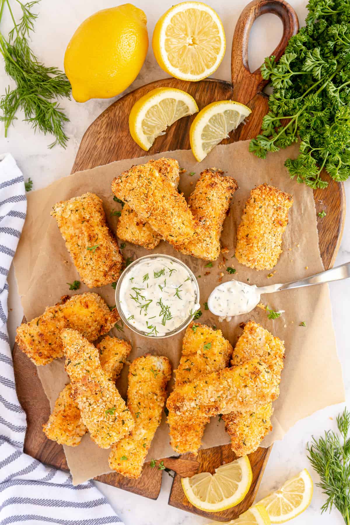 Fish sticks on a cutting board with tartar sauce and lemon wedges.