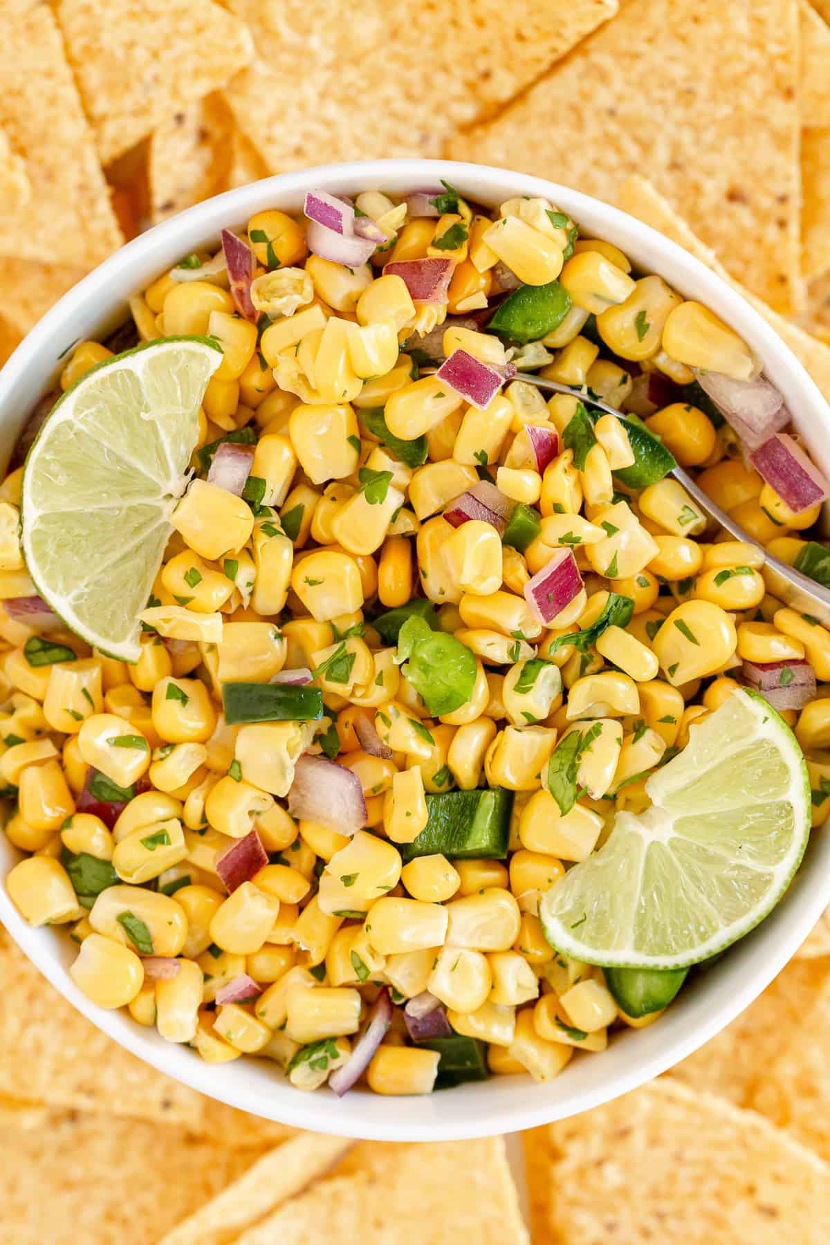 Corn salsa in a white bowl with lime wedges surrounded by tortilla chips.