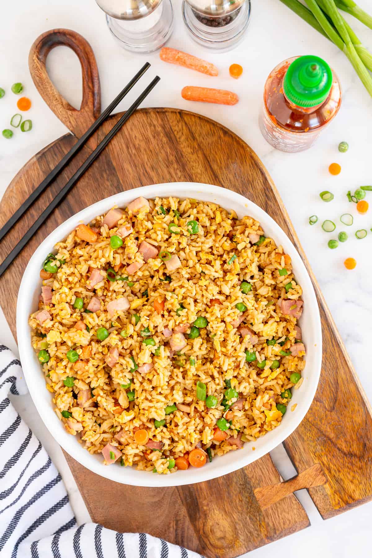 A bowl of Ham Fried Rice on a cutting board with a bottle of Sriracha behind it.