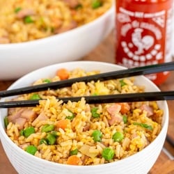A bowl of fried rice with a bottle Sriracha behind it.