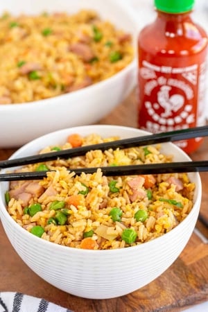 A bowl of fried rice with a bottle Sriracha behind it.