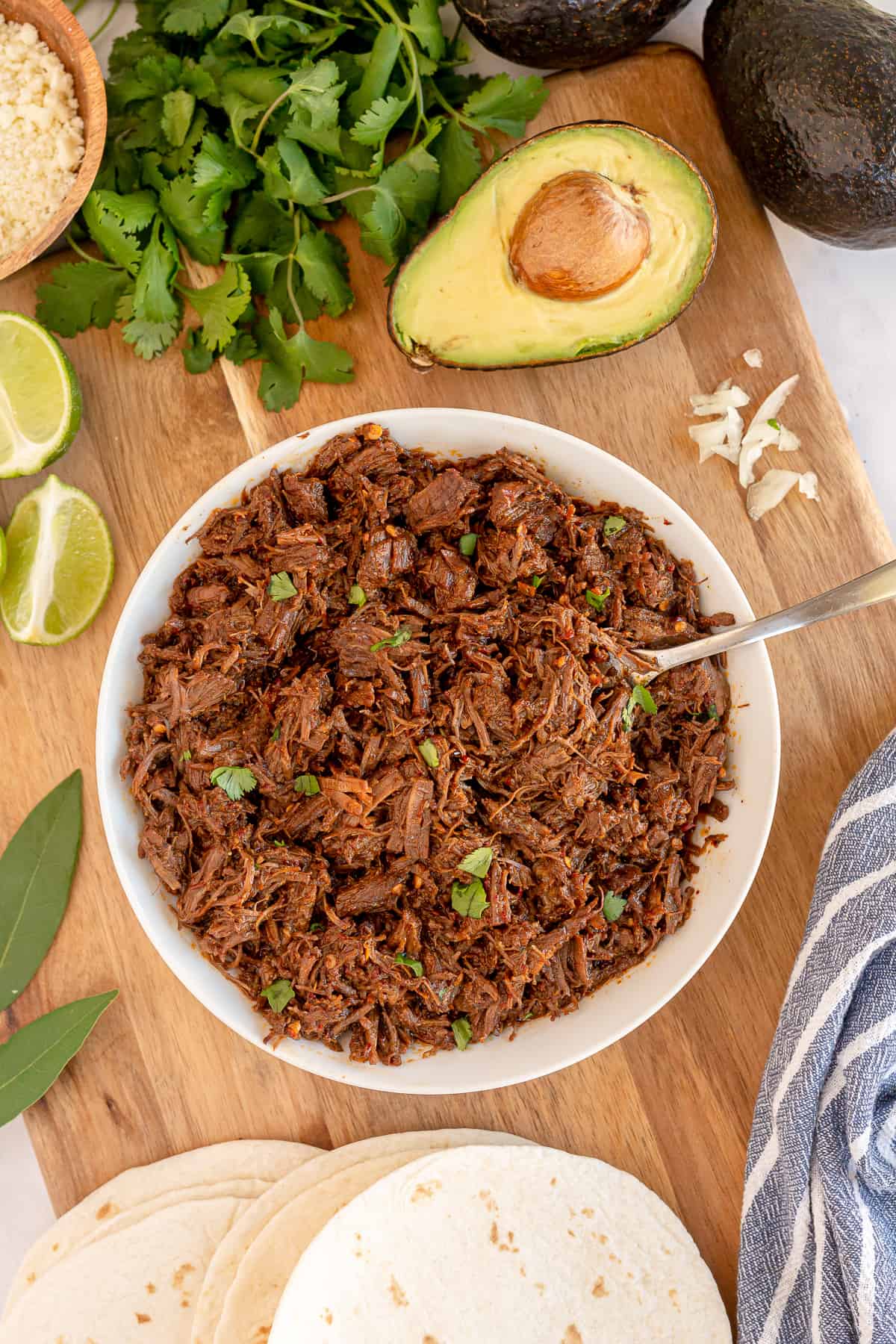 A bowl of Instant Pot Beef Barbacoa on a cutting board with other ingredients.