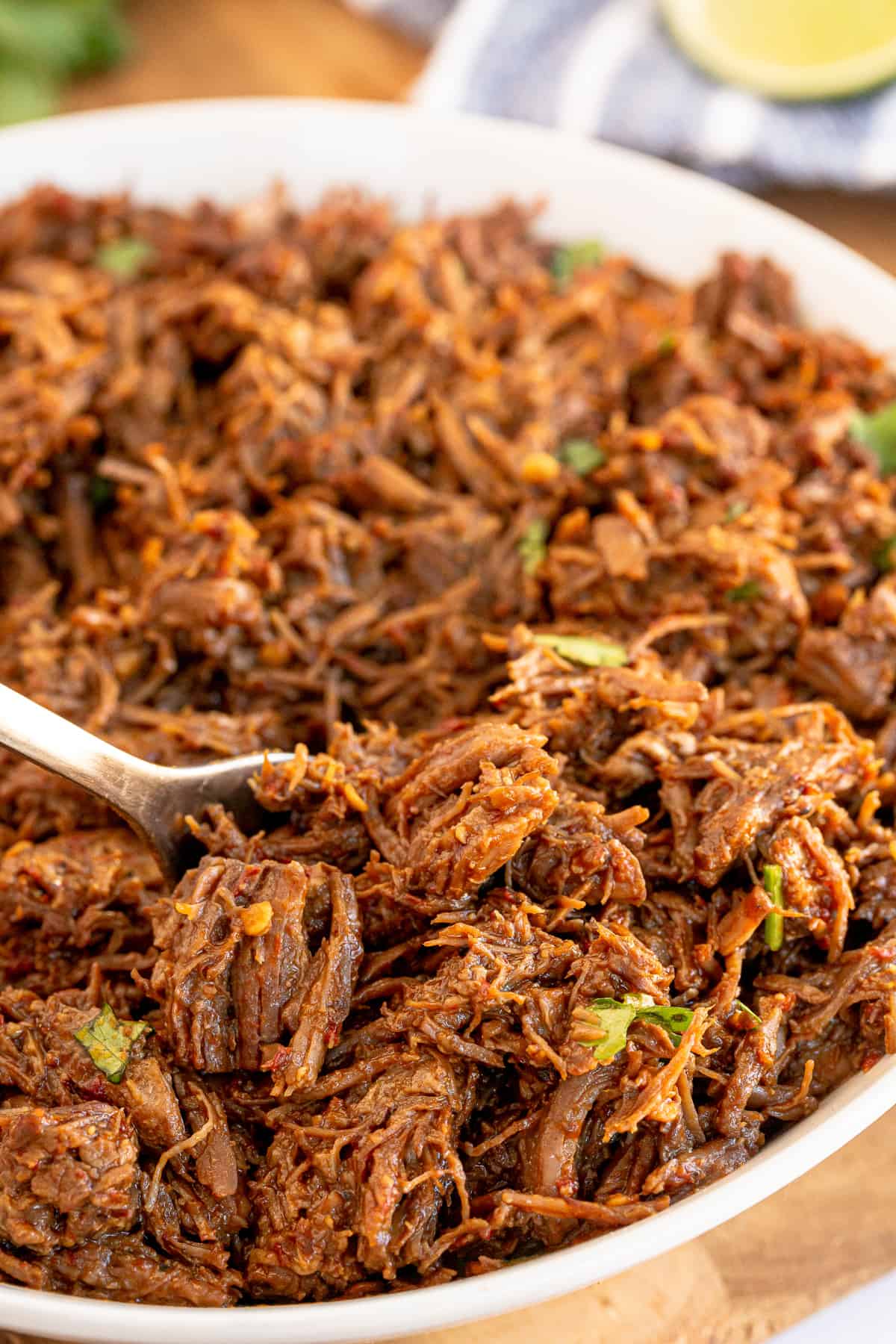 A fork digs into a mound of cooked, shredded beef barbacoa in a serving bowl.