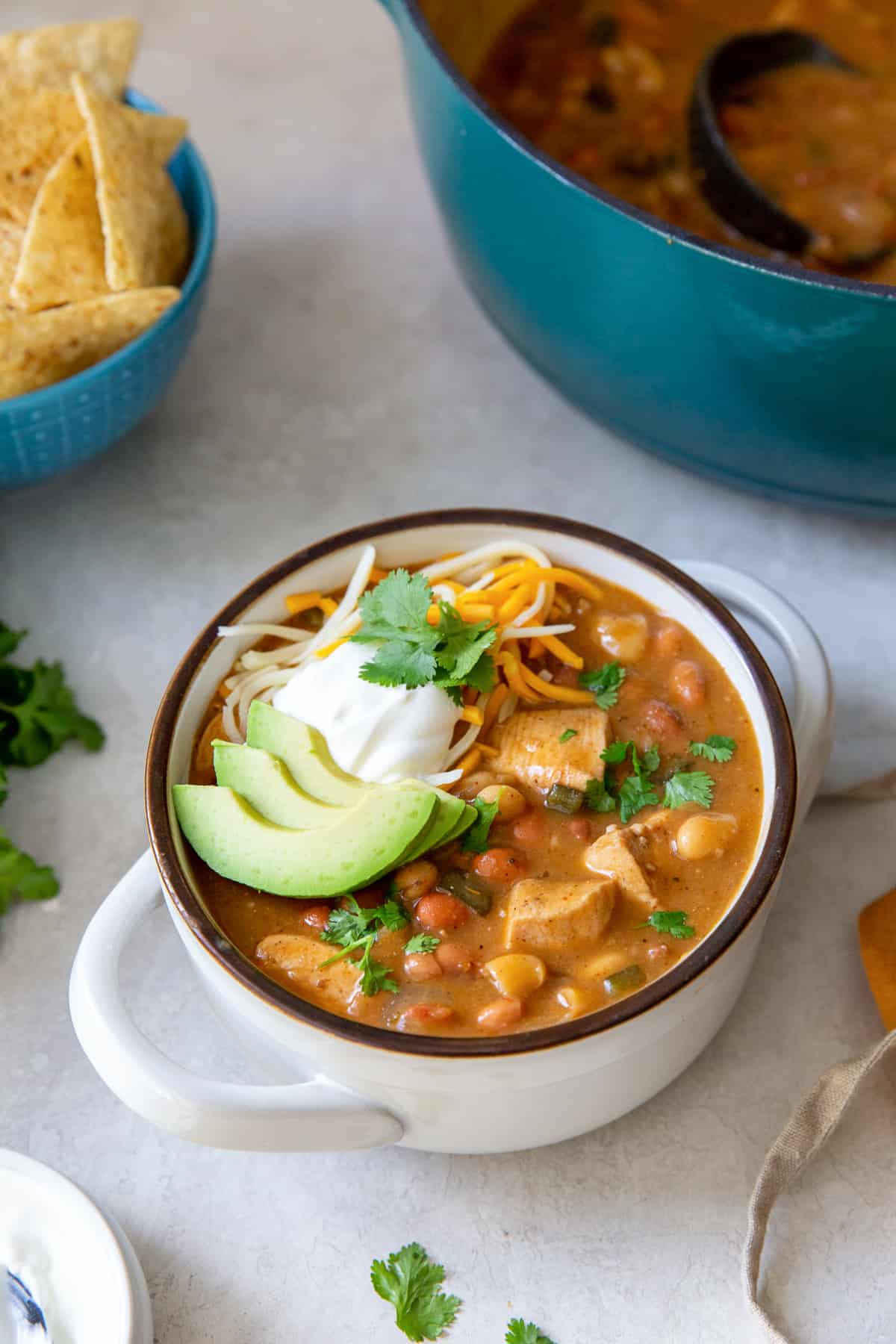 A bowl of chicken chili surrounded by a pot and tortilla chips.