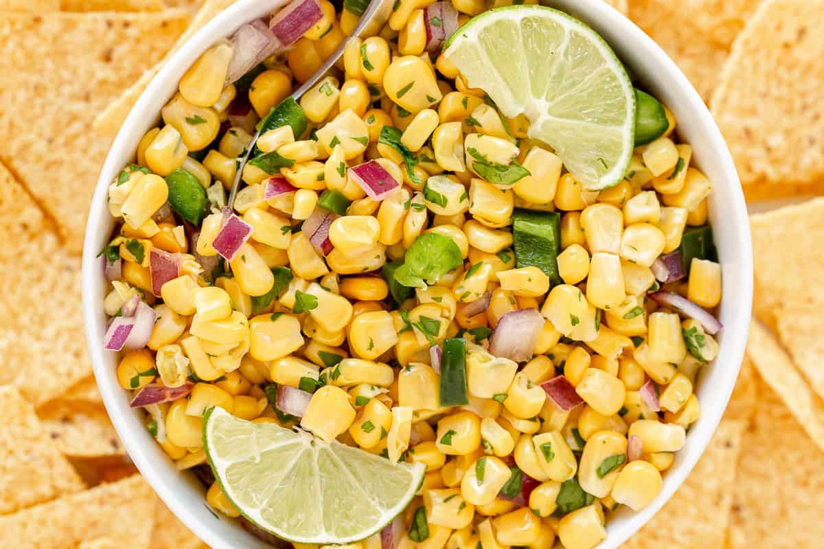 Chipotle copycat corn salsa with sweet corn, red onion, jalape&ntilde;o, cilantro, and lime wedges in a white bowl surrounded by tortilla chips.