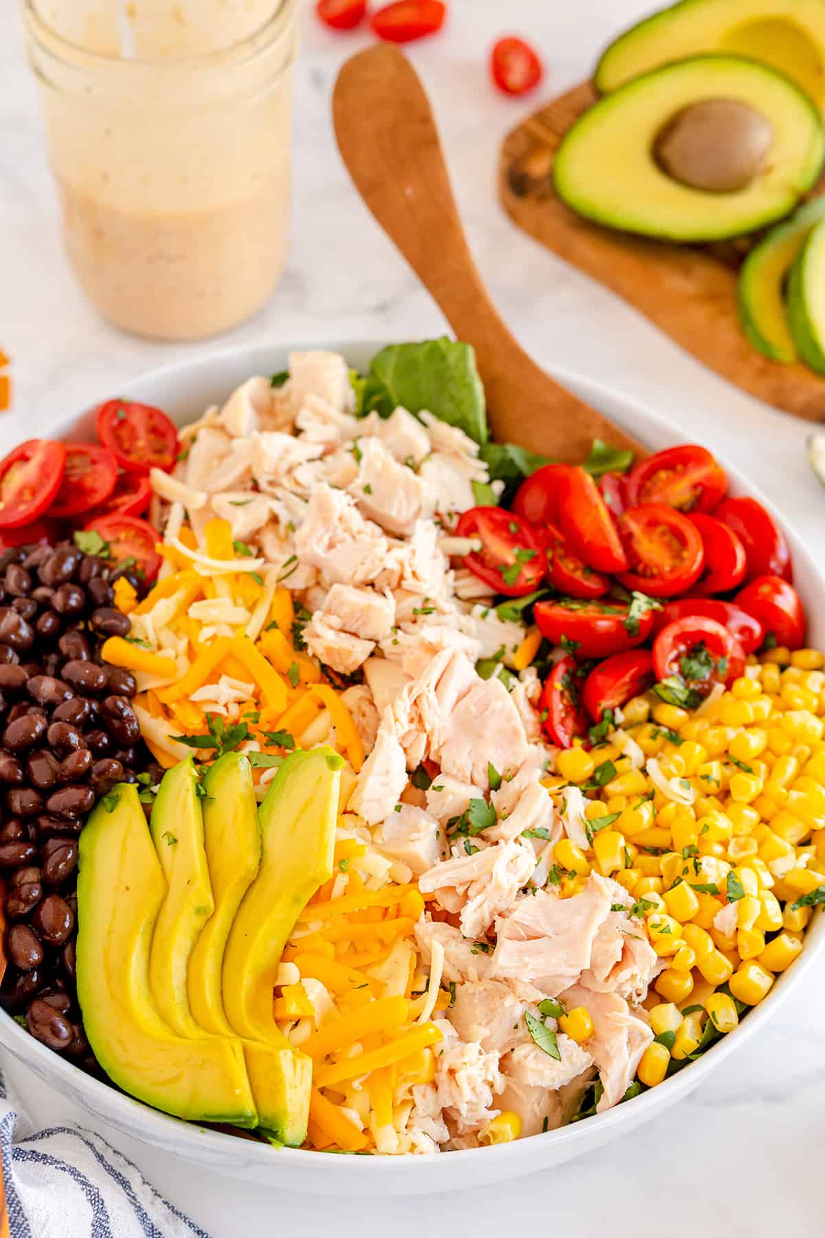 A Caesar salad with chicken, avocado, corn, and tomatoes in a bowl with a spoon.