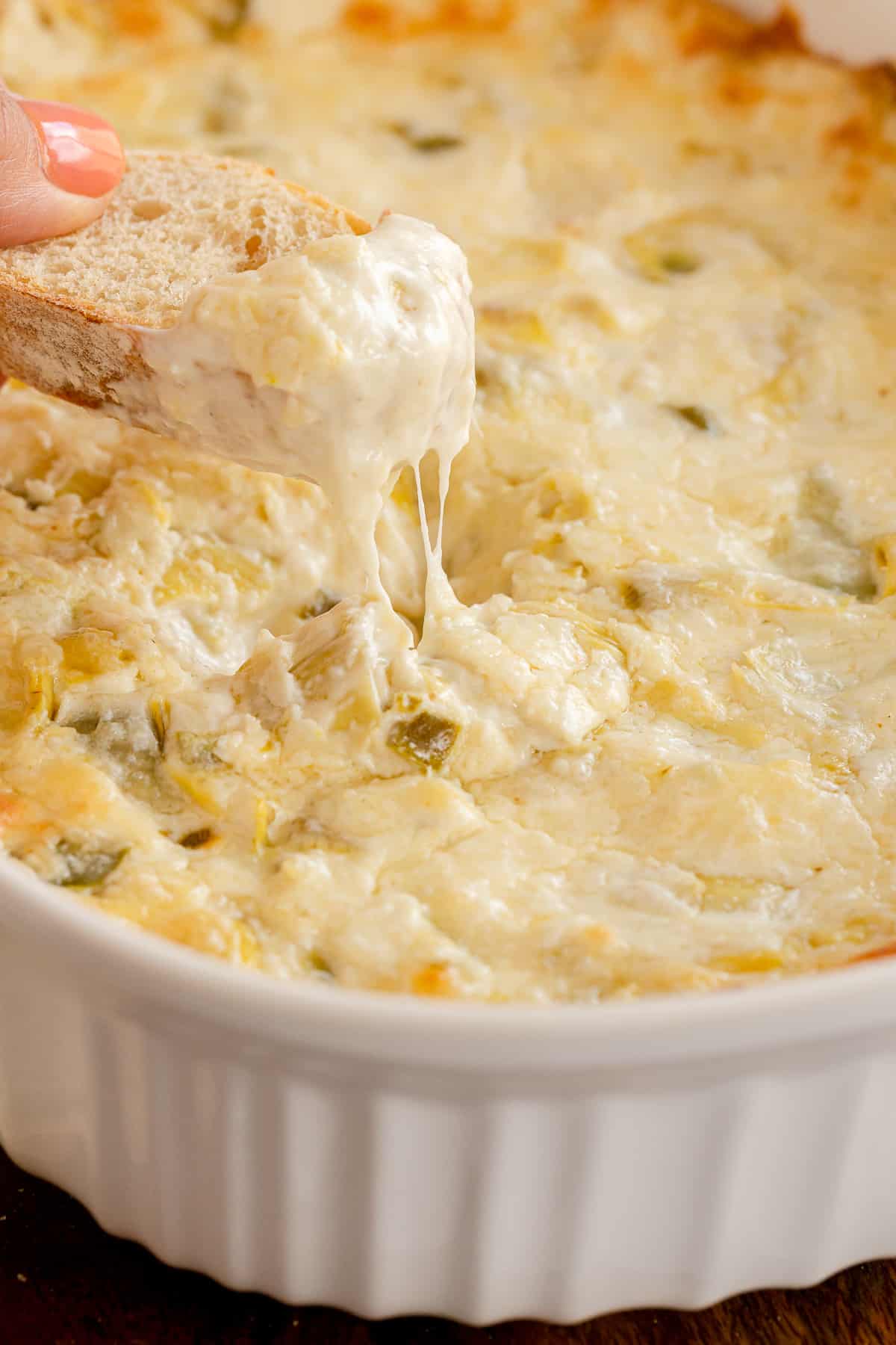 A hand dips a slice of bread into Cheesy Artichoke Dip.