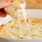 A hand dips a slice of bread into Cheesy Artichoke Dip.