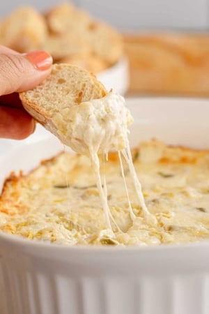 A hand dips a slice of bread into Cheesy Artichoke Dip.