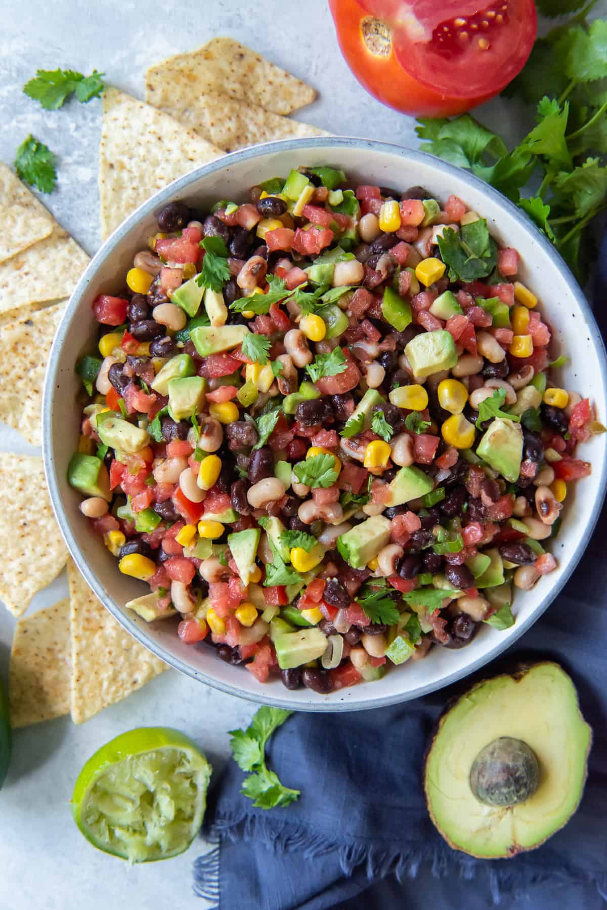 A bowl of Cowboy Caviar next to an avocado, tomato and a squeeze lime half.