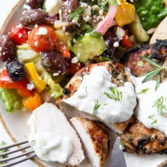Sliced Greek Chicken with Tzatziki on a plate with colorful Greek salad.
