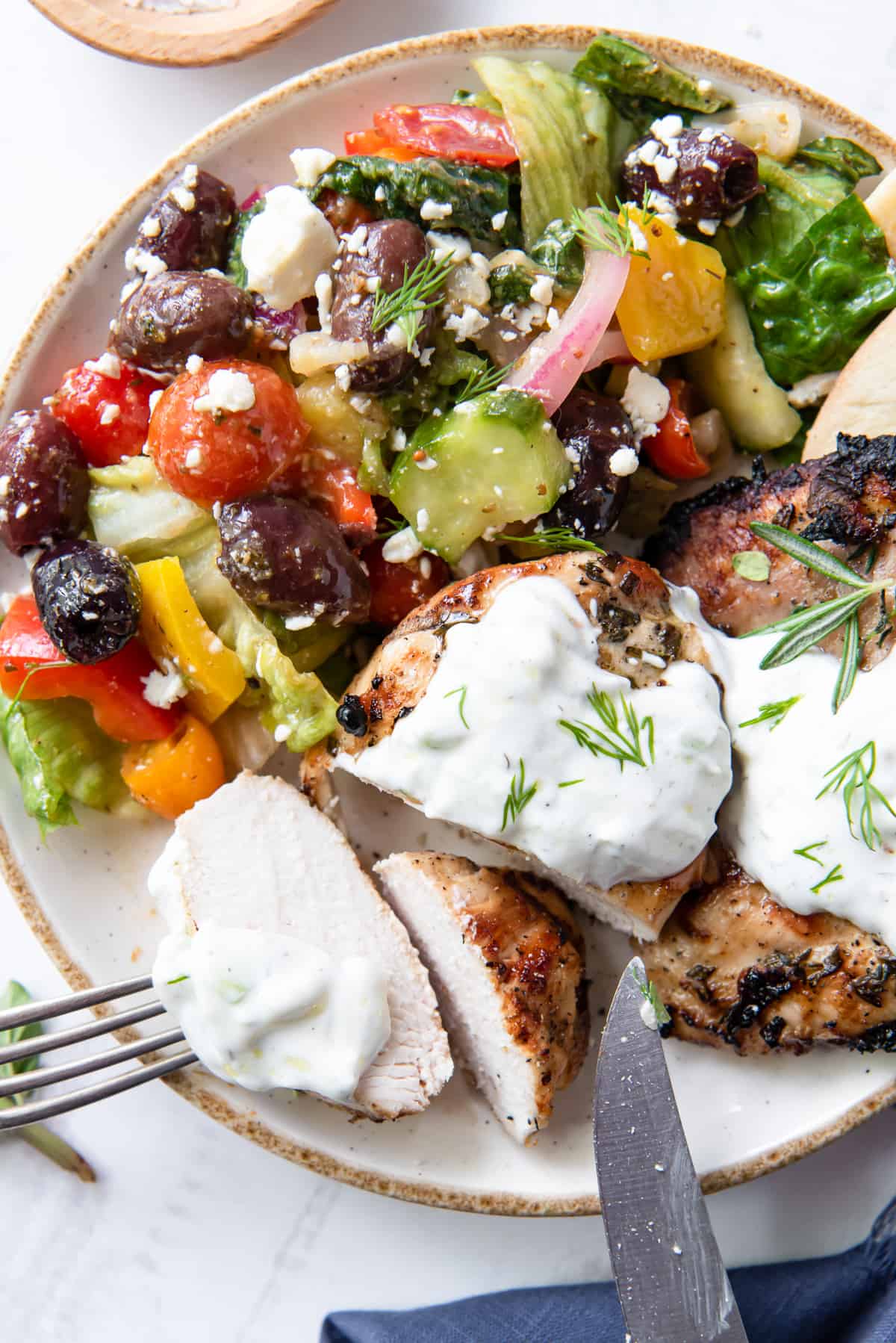 Sliced Greek Chicken with Tzatziki on a plate with colorful Greek salad.