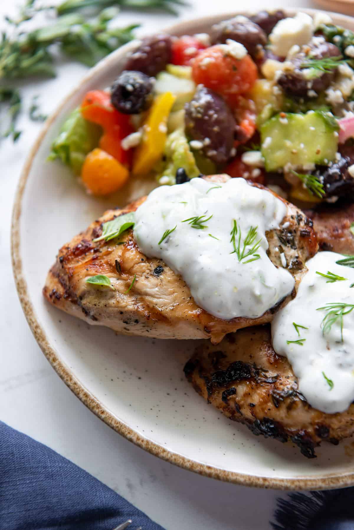 Two pieces of Grilled Greek Chicken topped with Tzatziki on a plate with salad.