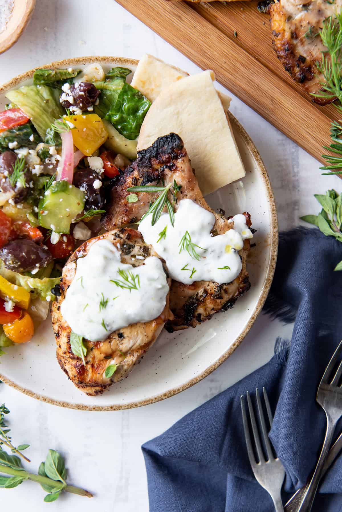Two pieces of Grilled Greek Chicken topped with Tzatziki on a plate with a colorful salad.