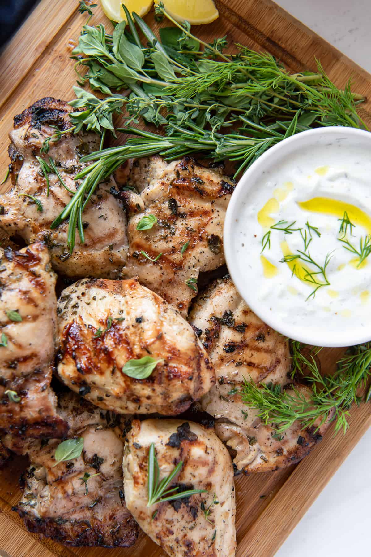 Grilled Greek Chicken on a cutting board with fresh herbs and Tzatziki Sauce.