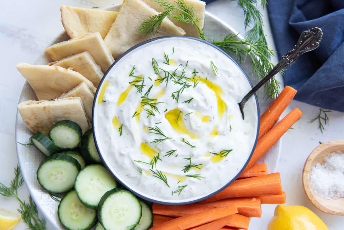 A bowl of Tzatziki on a platter with cucumber, carrots and pita.