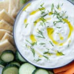 A bowl of Tzatziki on a plate with veggies and pita shot from over the top.