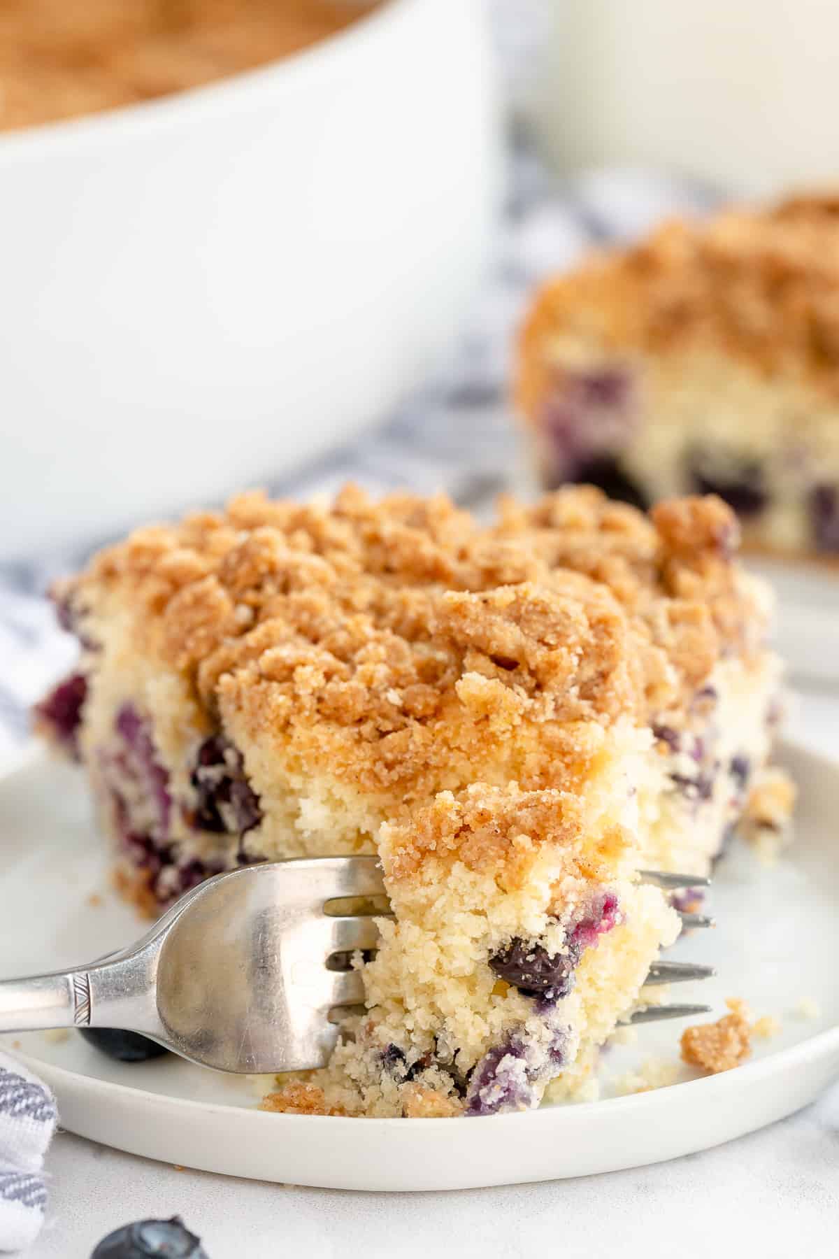 A fork pulls away a bite from a slice of Blueberry Crumb Cake.