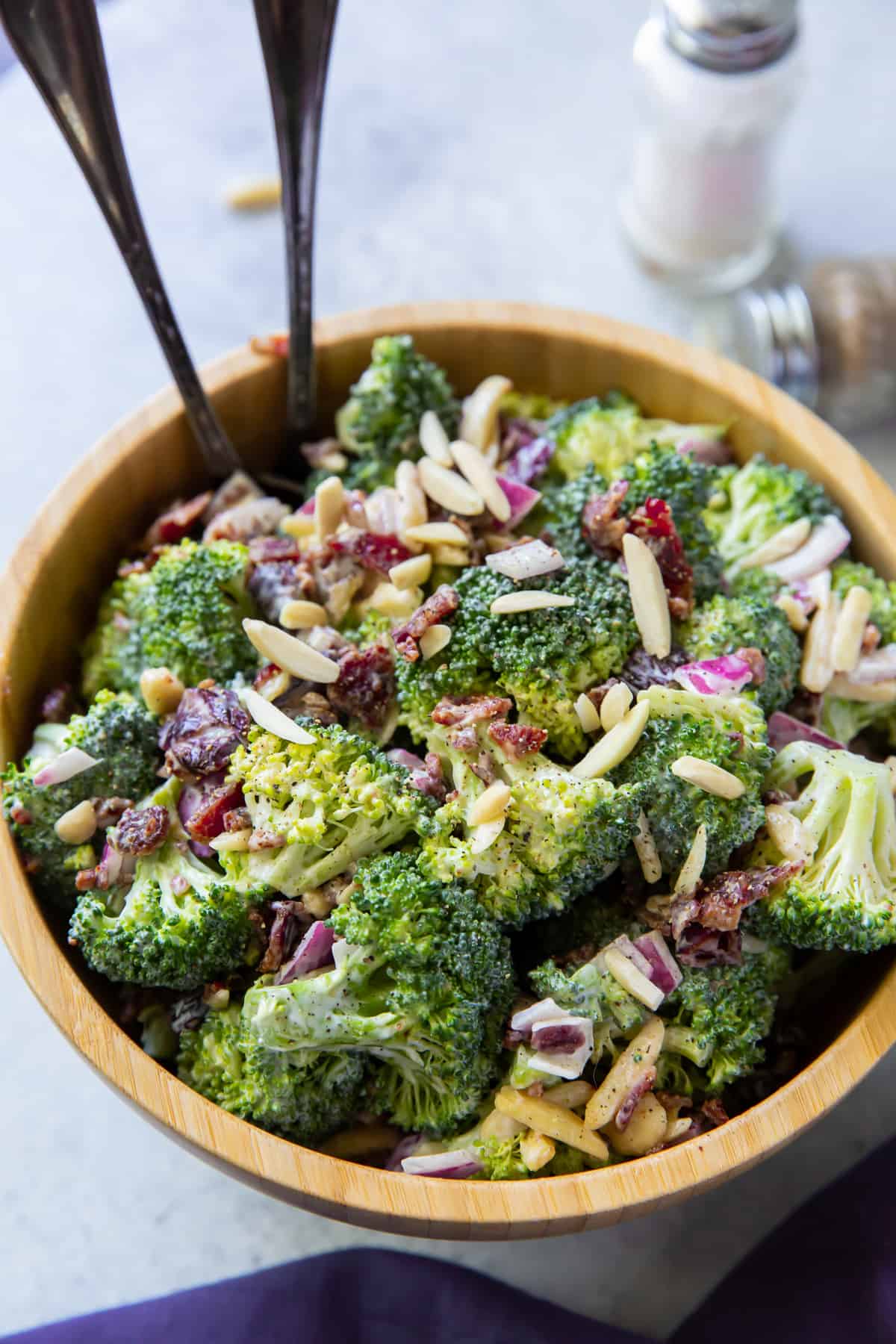 A wooden bowl filled with Broccoli Salad with two serving spoons sticking out of it.