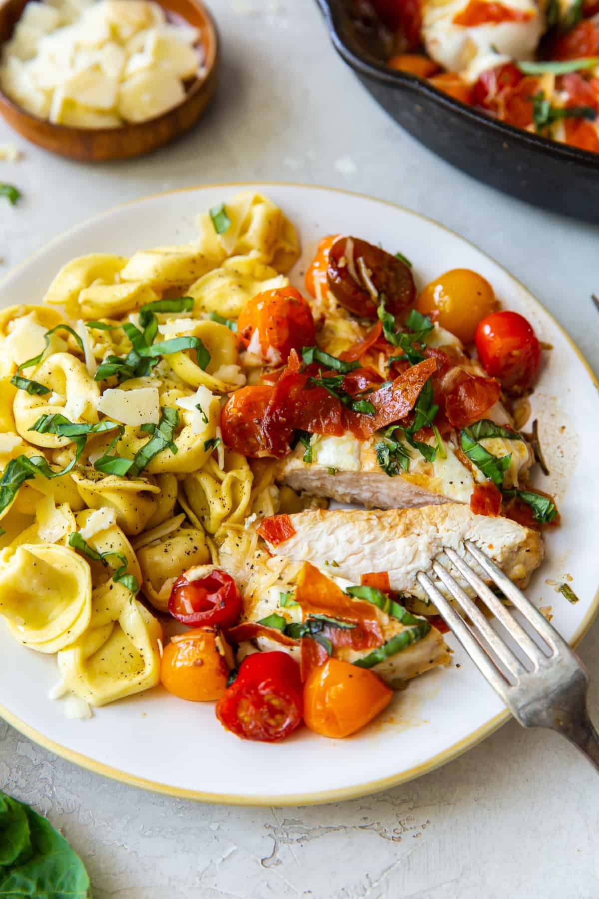 A fork presses into sliced chicken on a plate with tomatoes and tortellini.