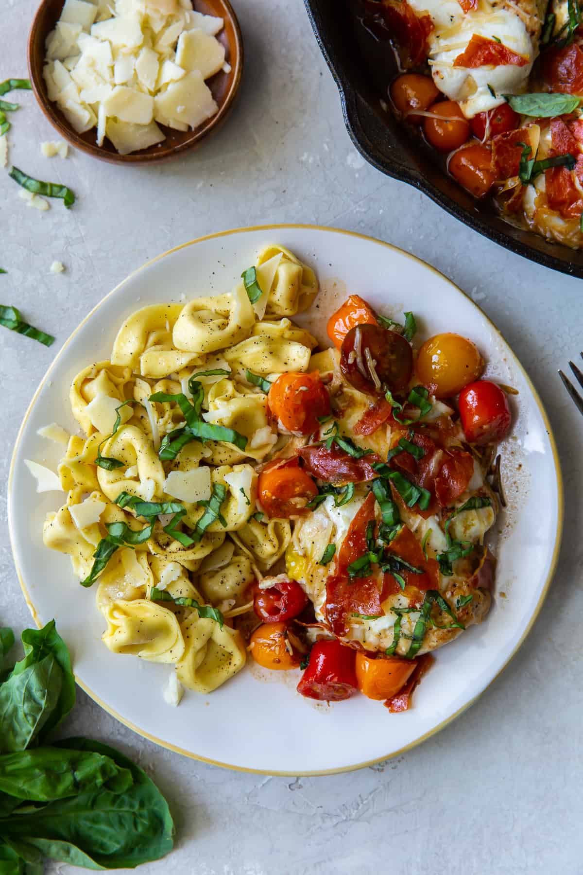 An over the top shot of a a plate of Caprese Chicken with tortellini next to a bowl of grated Parmesan.