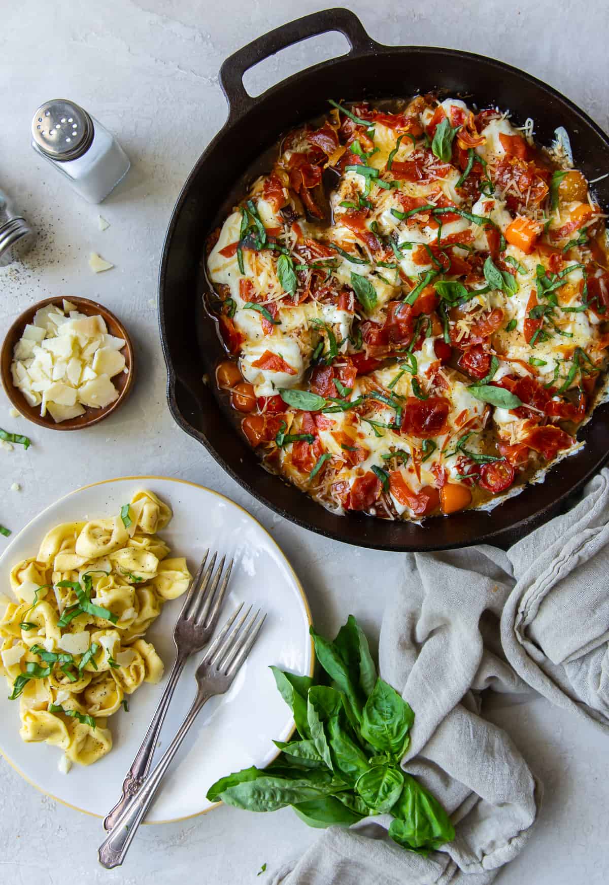 An over the top shot of Caprese Chicken in a skillet next to a plate of tortellini.