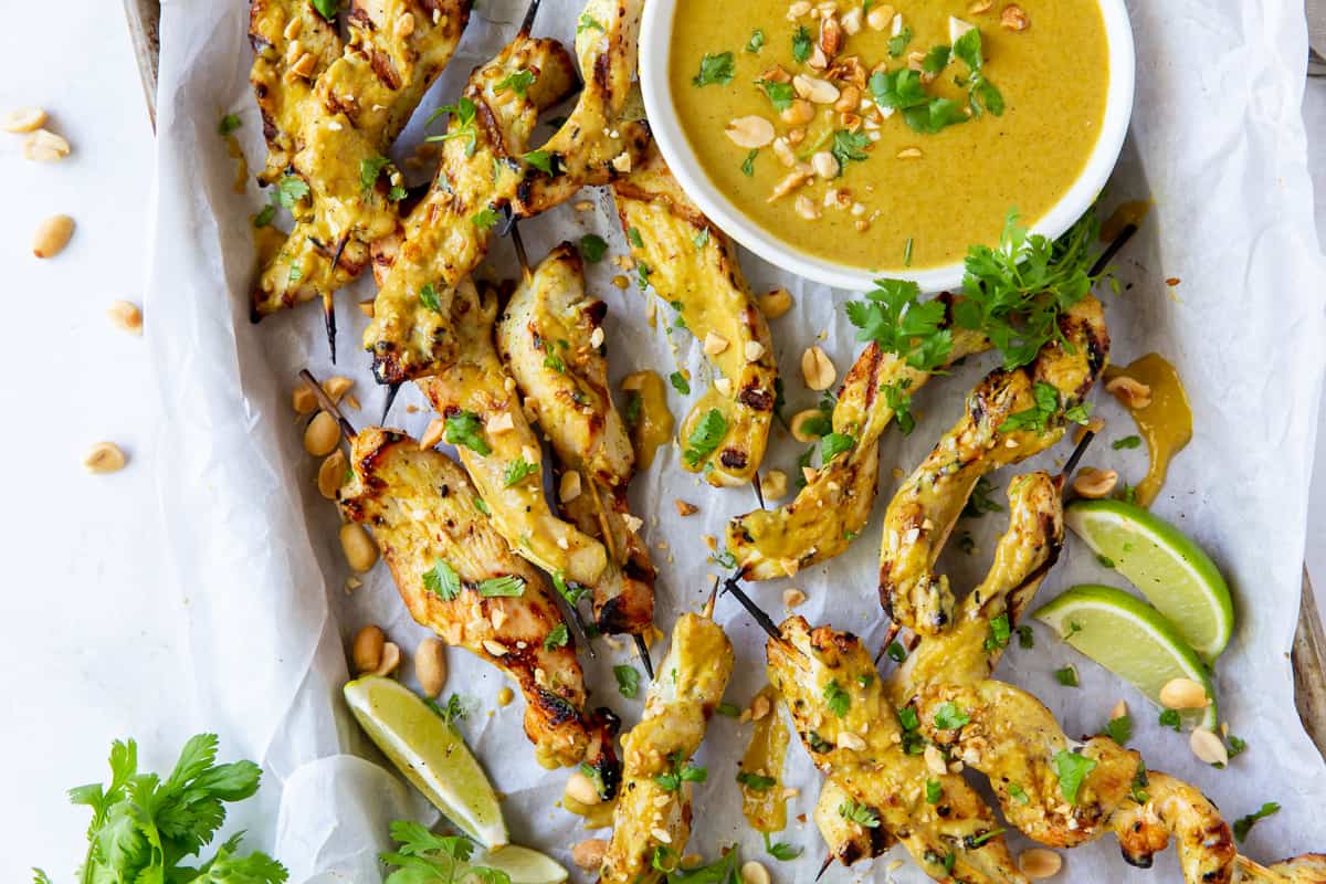 Grilled chicken satay skewers on a baking sheet with a bowl of peanut sauce.