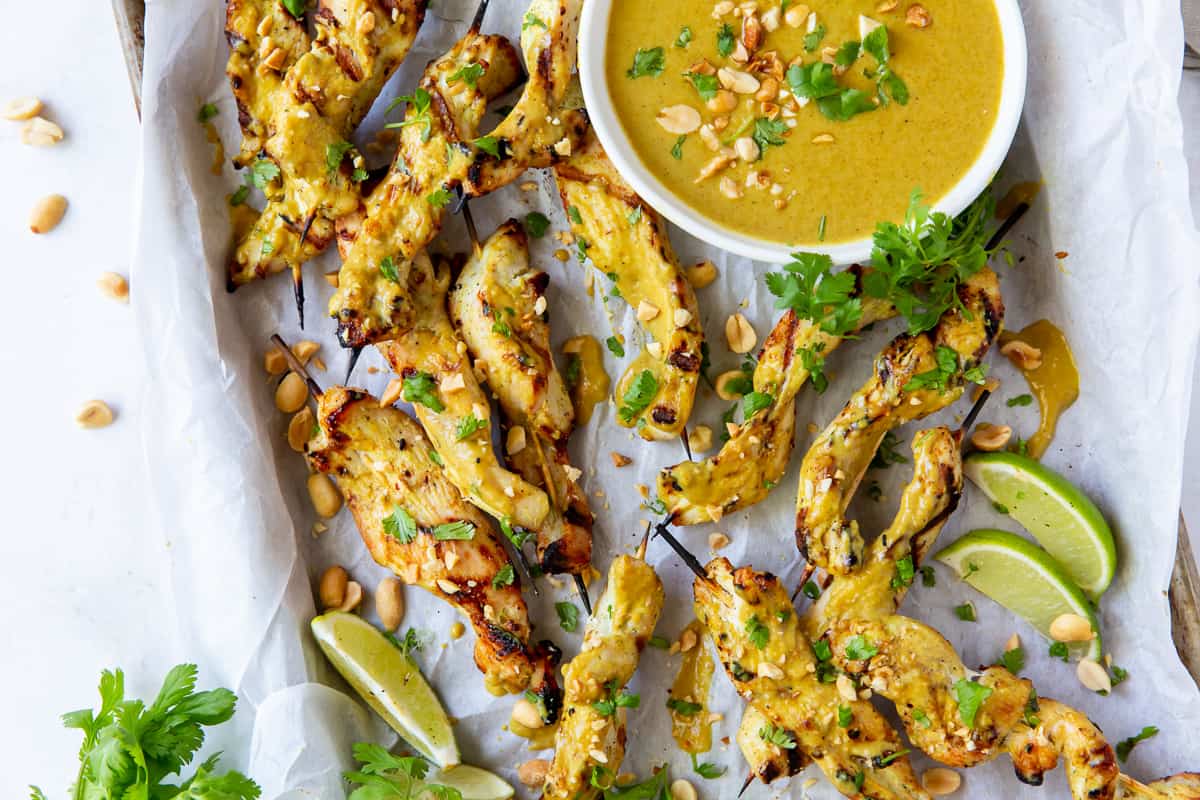 Grilled chicken satay skewers on a baking sheet with a bowl of peanut sauce.