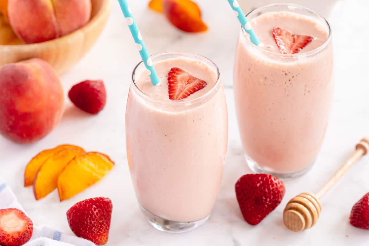 Two Strawberry Peach Smoothies with ingredients placed around them.