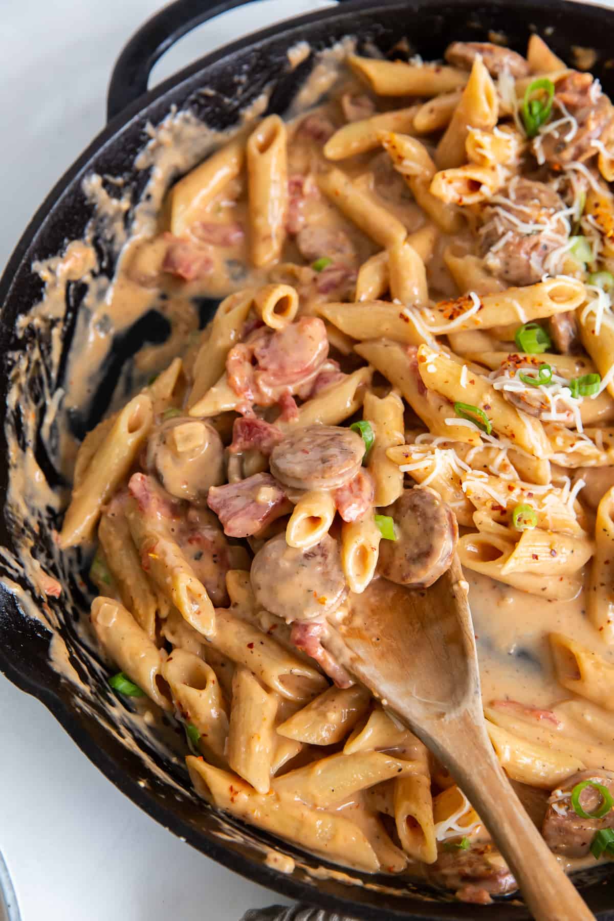 A wooden spoon resting in a skillet of sausage pasta.