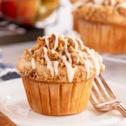 An apple muffin on a white plate in front of a crate of apples.