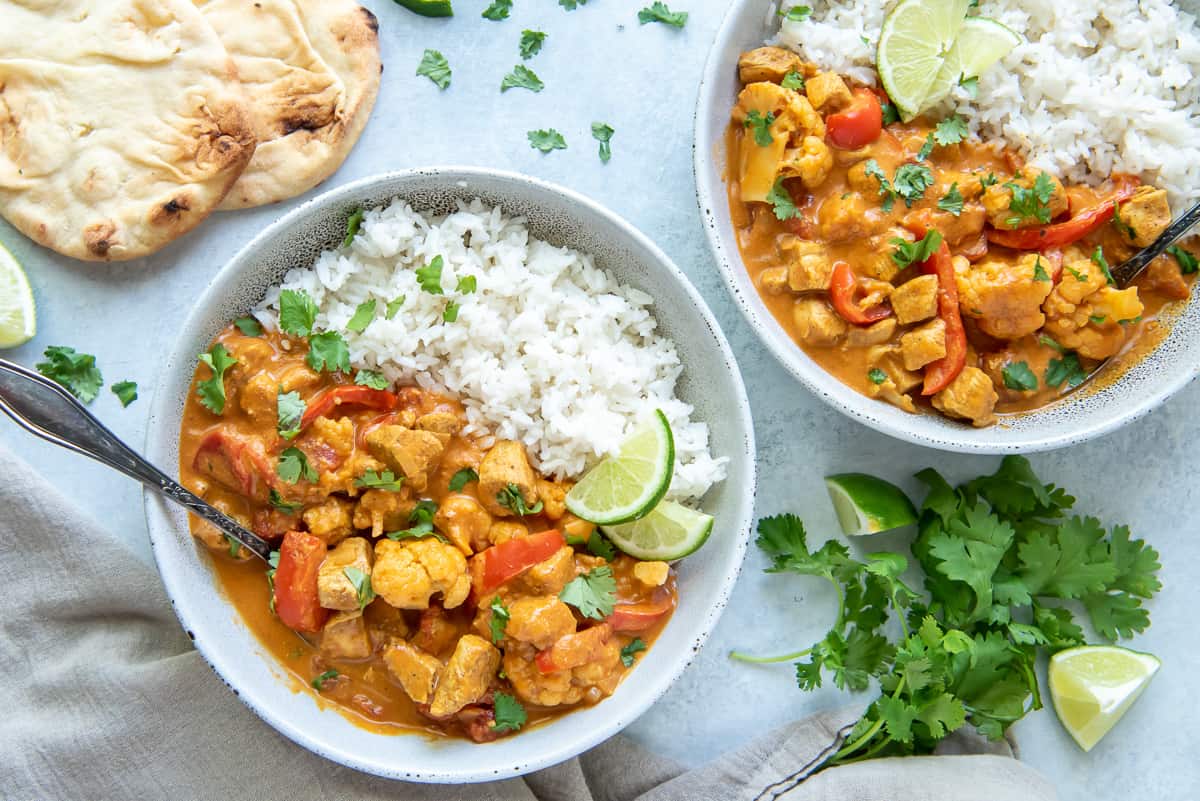 Two bowls of curry with chicken and rice next to pieces of naan and cilantro.