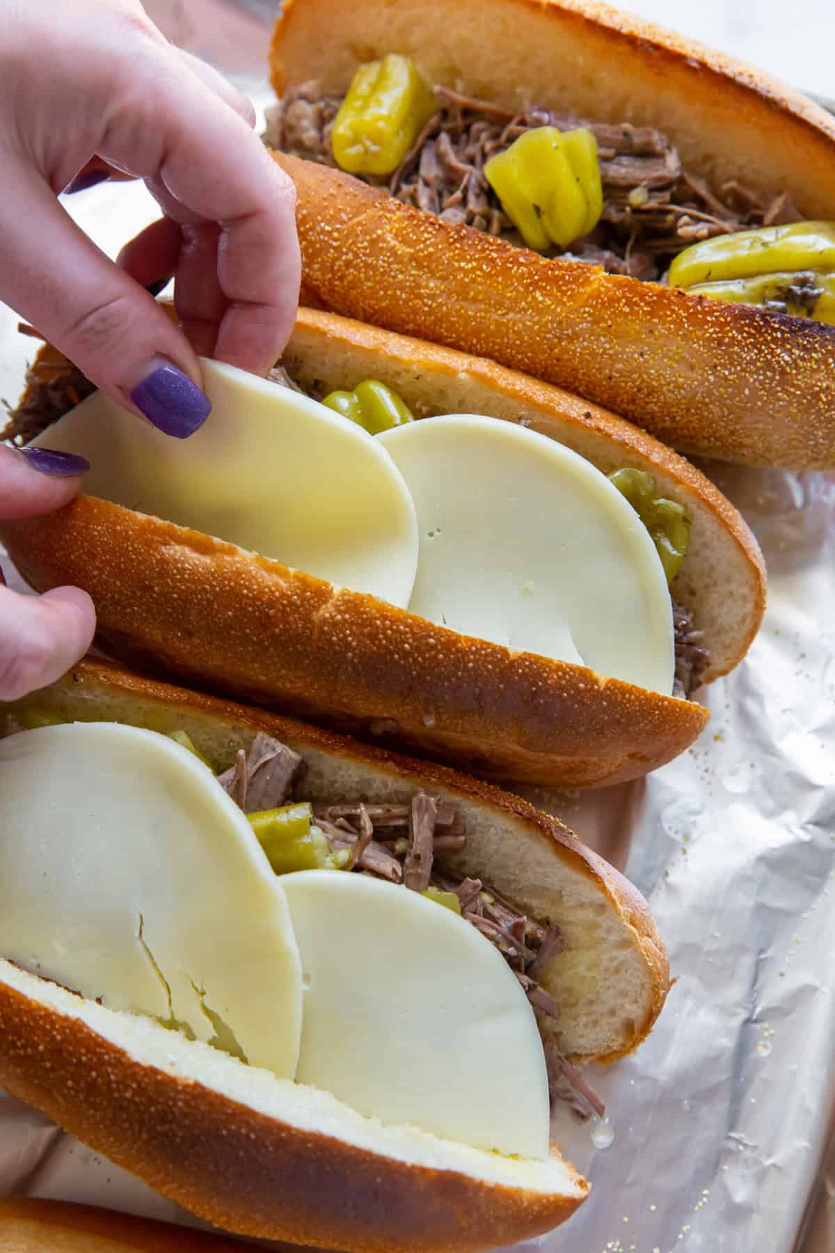 A hand placing sliced provolone cheese on top of Italian beef in sandwich rolls.